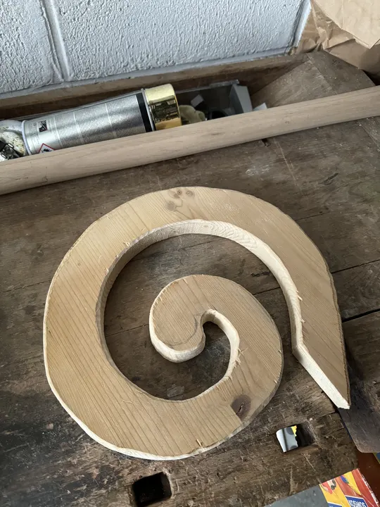 Unfertiger Kopf eines Hirtenstabes liegt in Form einer ausgesägten Holzspirale auf einer Werkbank.
