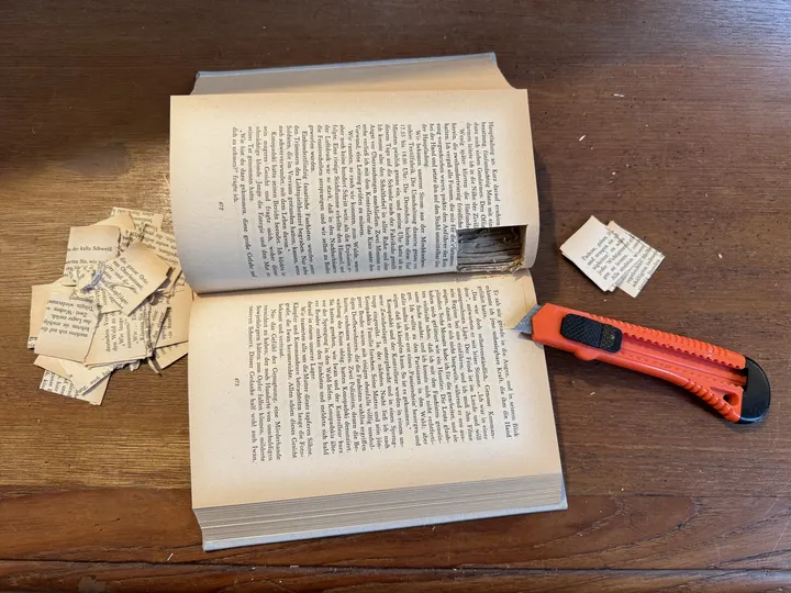 Aufgeklapptes Buch mit einem ausgeschnittenen Versteck für eine Kamera. Daneben ein Teppichmesser und Papierschnippsel