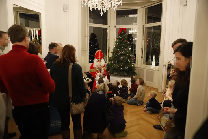 Nikolaus sitzt umringt von den sitzenden Kindern. Drumherum stehen Erwachsene.