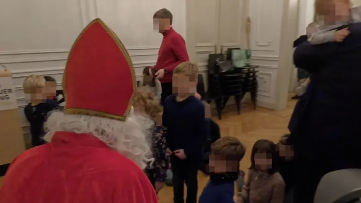 Rücken des Nikolaus mit den ihn anschauenden Kindern. Im Hintergrund geht ein blonder Mann mit dickem Bauch unter einem roten Pullover.