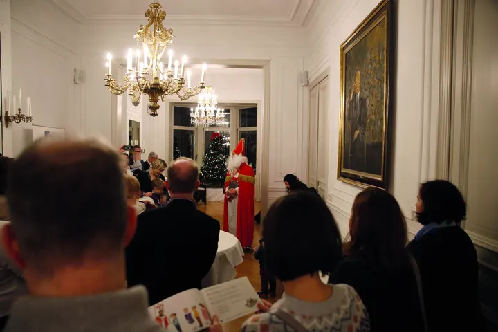 Weitwinklige Aufnahme des festlichen Saals mit Kronleuchter und der singenden Feiergesellschaft von ca. 40 klassisch gekleideten Erwachsenen und 20 rausgeputzten Kindern. An der rechten Wand hängt Sutherlands Porträt von Konrad Adenauer (nachdenklich, auf dunklem Hintergrund)